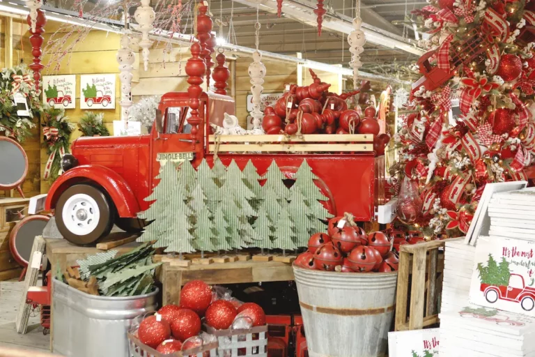 A festive display features a red vintage truck loaded with red decorations among Christmas trees, ornaments, and holiday-themed items in a store, capturing the spirit of fowl and flame.
