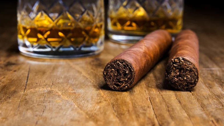 Two cigars rest on a wooden surface with two glasses of amber-colored whiskey in the background
