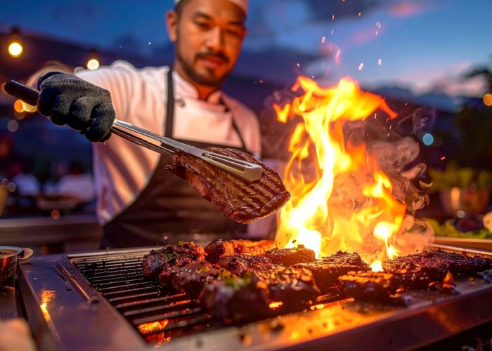 A chef grilling steaks over an open flame on an outdoor grill at dusk, with flames rising as he uses tongs to turn a piece of meat.