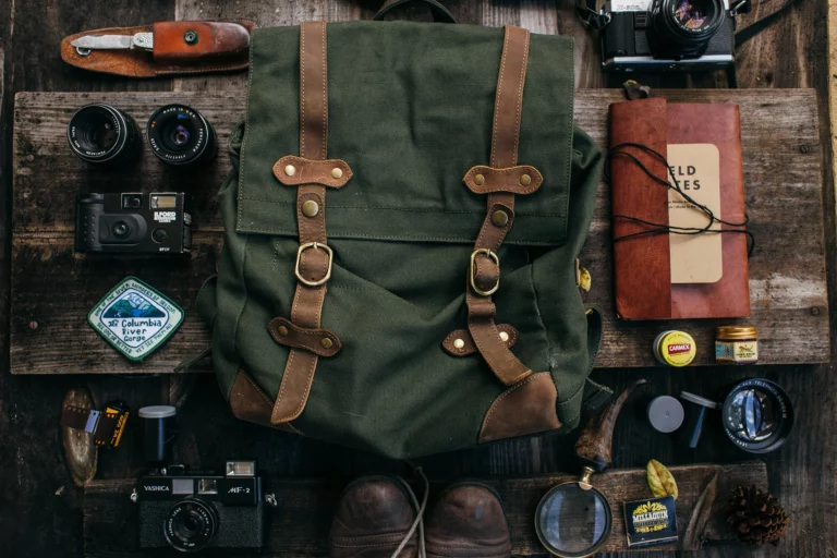 A green canvas backpack surrounded by cameras, a notebook, film, a map patch, and hiking boots rests on a wooden surface—ready for adventure with a fowl and flame spirit.