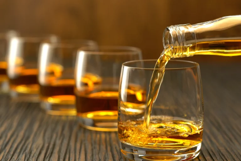 A close-up of whiskey being poured from a bottle into a glass, with three more filled glasses lined up in the background on a wooden surface.