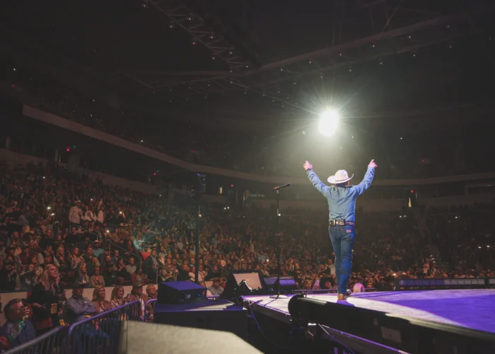 A performer in a hat stands on stage with arms raised, facing a large cheering audience in a well-lit indoor arena.