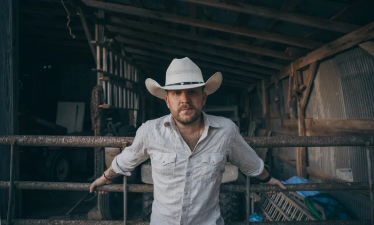 A man wearing a white cowboy hat and light shirt leans on a metal fence inside a rustic barn.